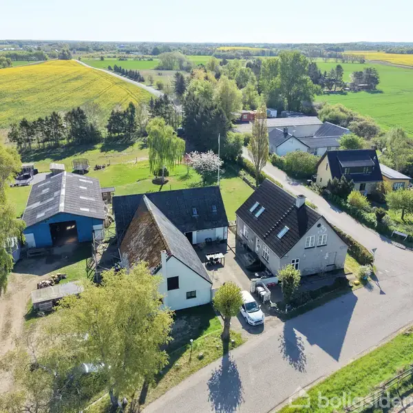 Hus i landligt område, omgivet af grønne marker og træer, med flere bygninger og en parkeret bil på en vej. Boligerne er placeret tæt sammen med et landligt baggrundspanorama.