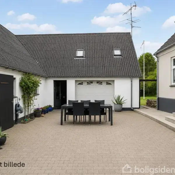 En gårdhave med sorte udendørsmøbler står på en flisebelagt terrasse mellem to huse. Der er døre, vinduer og planter langs bygningerne. Tekst: "AI genereret billede".