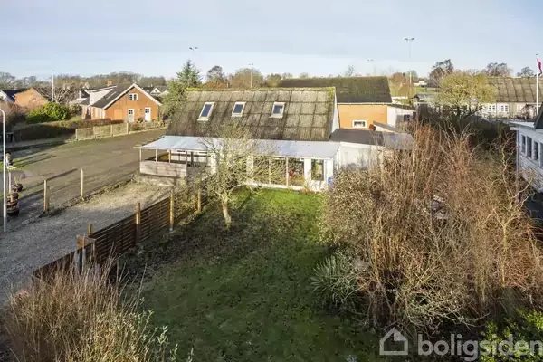 Et hus med skråtag ses i en have med buske og træer. Baggrunden viser flere huse og en sti.