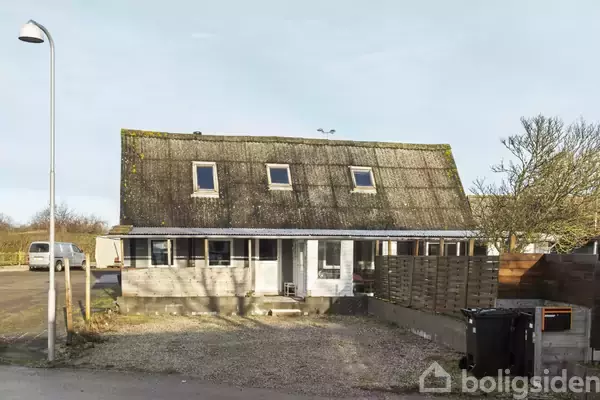 Et ældre hus med skråt tag og fem vinduer står i en lille have med grusbelægning. En lygtepæl og affaldscontainere ses i nærheden.