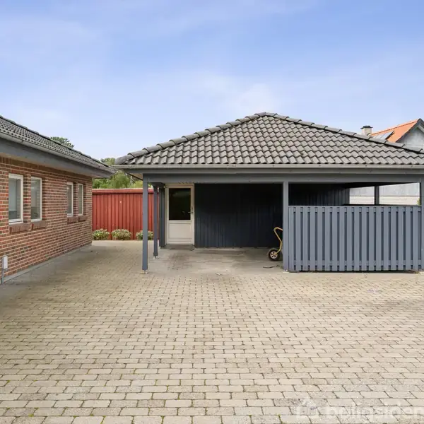 Carport med gråt tag står i indkørsel belagt med grå fliser. Til venstre ses en muret bygning med rødt murværk og hvide vinduer. Grønt buskads bagved.