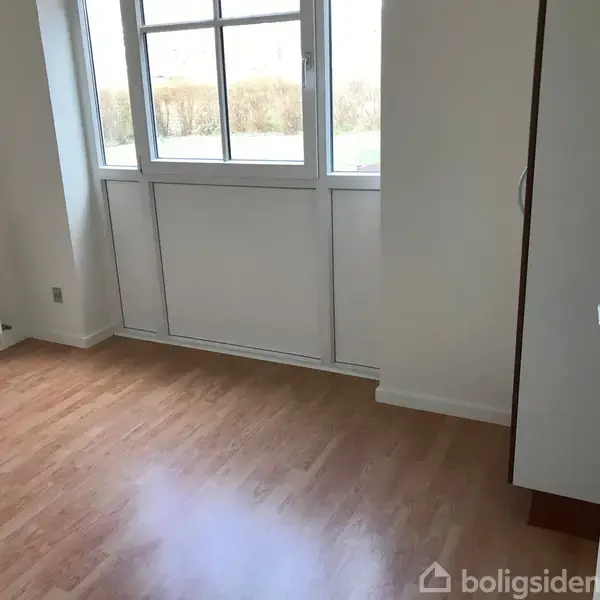 Et lyst, tomt rum med trægulv og et stort vindue, der slipper naturligt lys ind. Døren står åben, og der er et radiator under vinduet.