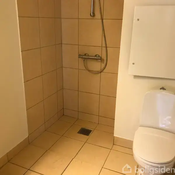 Toilet placeret ved siden af et bruseområde uden adskillelse, med beige fliser på gulvet og væggene. Et skab hænger på væggen over toilettet. Ingen tekst til stede.