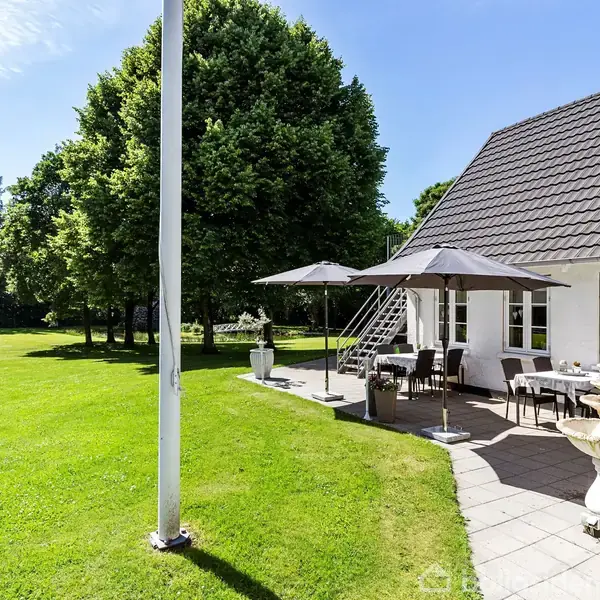 Hvid hus med sort tag og terrasse fyldt med borde, stole og parasoller i skyggefuld have med græsplæne og træer. Flagstang ved siden af huset.