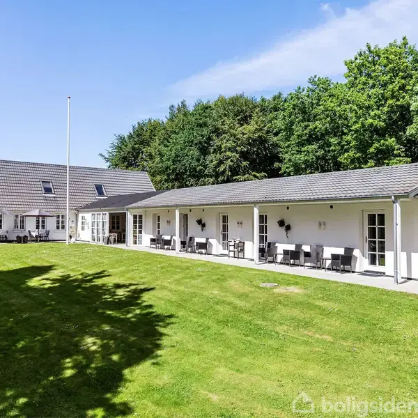 Et moderne, hvidt hus med terrasse i solrigt vejr, omkranset af en stor, velplejet græsplæne og tæt bagvedliggende skov.