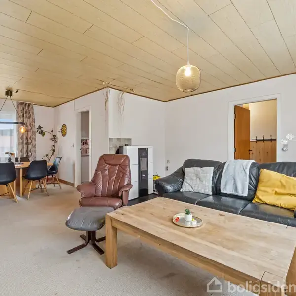 En stue med en sort sofa, et træbord og en lænestol. Spisestuen i baggrunden har et spisebord omgivet af stole. Rummet er lyst med vinduer og neutralt interiør.