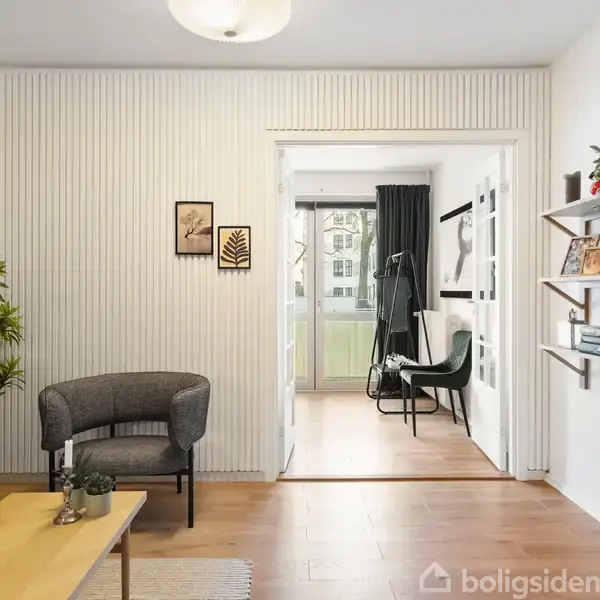 Stue med sofa og lænestol, hvide vægge og træpaneler. Reoler med bøger og planter. Åben dør til lys balkon med stole. Mønstrede billeder på væggen.