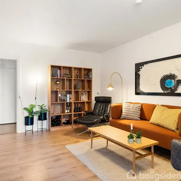 En stue med en brun sofa og sofabord i træ. En lænestol står ved siden af en reol fyldt med bøger og pynt. Væggene er lyse, og der hænger et stort kunstværk over sofaen.