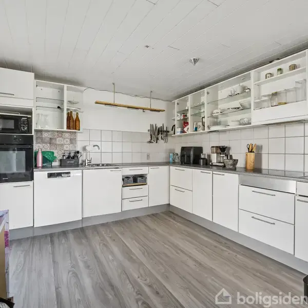 Hvidt køkken med L-formet layout og trælaminatgulv. Skabene har glaslåger og stå på væggen sammen med en indbygget ovn og mikrobølgeovn. Diverse køkkenredskaber og apparater er placeret på bordene.
