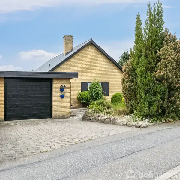 Gulstenshus står stille på en boligvej, flankeret af frodig buskads og små træer, med en sort garageport og en brolagt indkørsel.