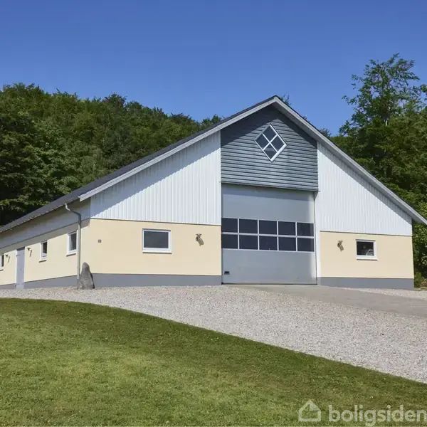 En stor, moderne ladebygning står tom på en grussti; omgivet af græs og tæt skov. Bygningen har en stor dør, to mindre vinduer og et kvadratisk gavlvindue.