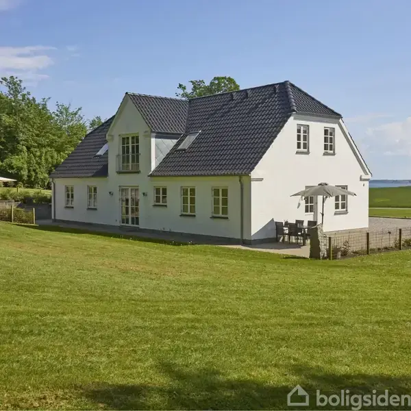 Hus med hvid facade og blå tagsten står på en grøn græsplæne. Solsenge og parasoller ses ved siden af, omgivet af træer og udsigt over havet i baggrunden.