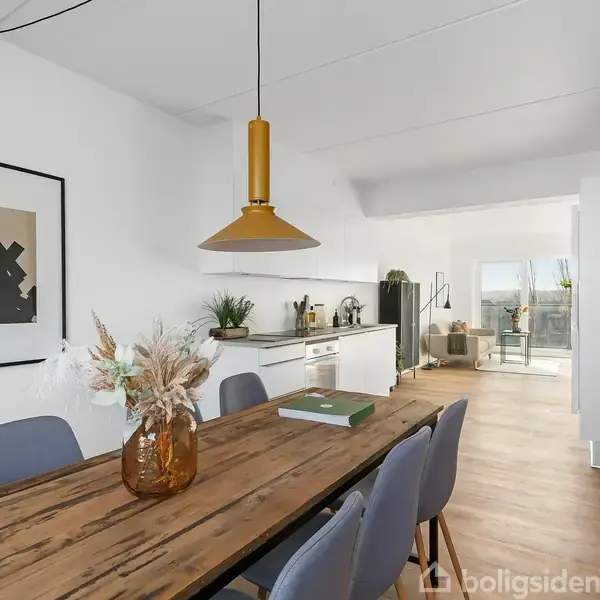 En spisebord med blå stole står i en lys spisestue. Over bordet hænger en gul lampe. I baggrunden ses et åbent køkken med hvide skabe og en balkon.