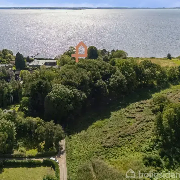 Hus midt i skovområde, markeret med orange ikon, nær vandkanten. Omringet af grønne træer og åbne marker. Vandet strækker sig mod horisonten under en klar himmel.