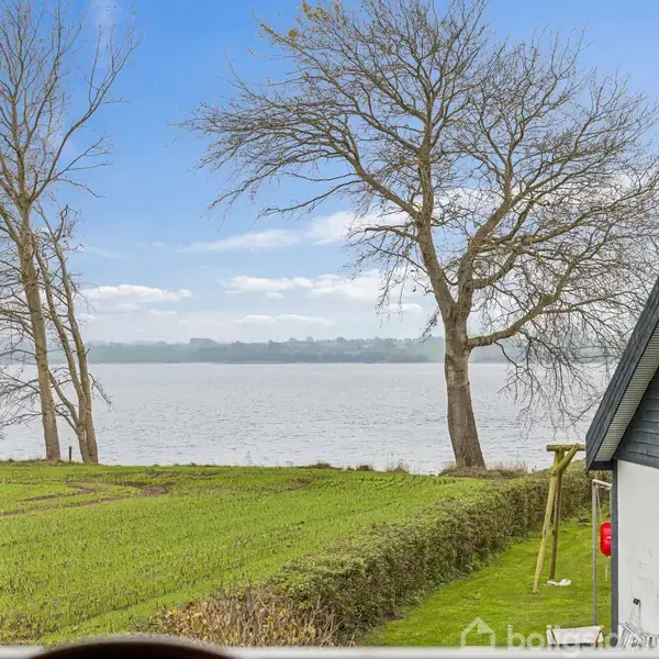 Et landskab set fra et vindue med en udsigt over en sø. Der er træer nær vandet, og et hus ses delvist til højre. Himmel er blå og klar.