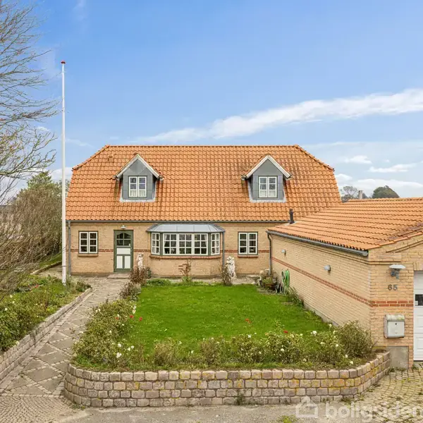 En murstensvilla med rødt tegltag har to kviste. Haven har en lille græsplæne og indkørsel. Der står nummer 65 på garagevæggen. Huset har flere vinduer og en hoveddør.