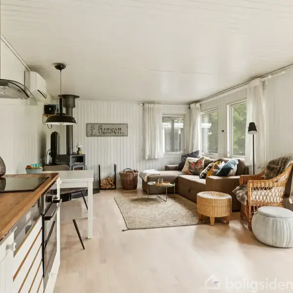 Et moderne køkken støder op til en stue med trægulve og store vinduer. En sofa, stole, og et skilt med teksten “Hansen” skaber en indbydende atmosfære.