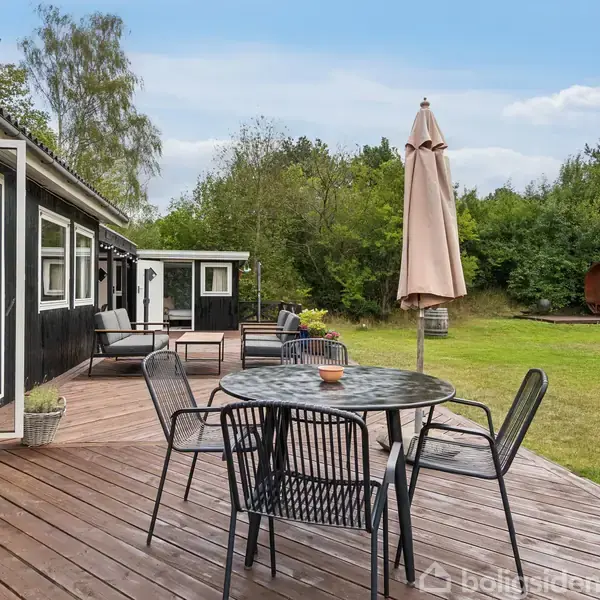 En terrasse med et rundt bord og fire stole, hvoraf den ene har en lukket parasol ved siden af. Bagved ses et sortmalet hus og en have med græsplæne og træer.