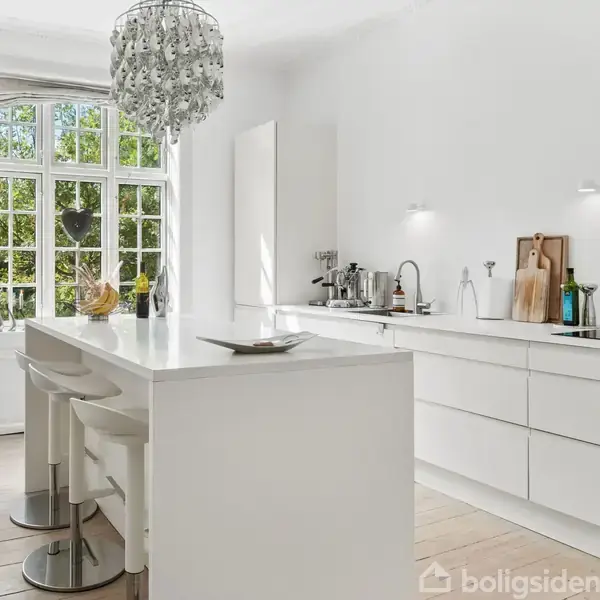 Et lyst køkken med en hvid ø og barstole i forgrunden; store vinduer bagved slipper naturligt lys ind. Der er køkkenredskaber og en vask på den indbyggede bordplade.