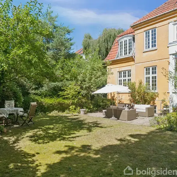 Gul bygning med hvide vinduer omgivet af en grøn have. Havemøbler og parasol står i solen ved siden af huset; et havesæt står i skyggen til venstre.