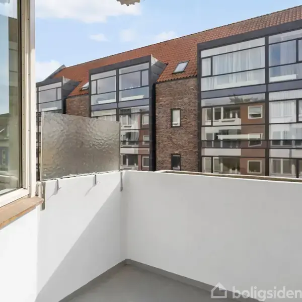 En lille, tom balkon med hvide vægge vender ud mod en moderne bygning med rødt tegltag og store vinduer. Sollys stråler ind, skaber en lys atmosfære.
