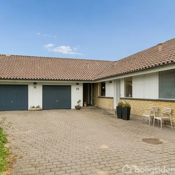 Et gult murstenshus med tegltag har to garageporte og en flisebelagt indkørsel. Huset er omgivet af grønne buske. Nummeret "75" ses ved hoveddøren, og tre stole står udenfor.
