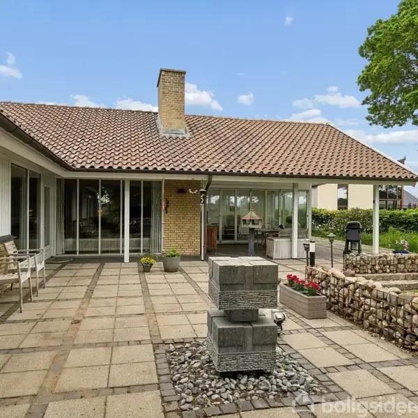 Terrasse med flisebelægning, centreret om en lille vandfontæne på sten, omgivet af en gul murstensbygning med store vinduer. I baggrunden ses en grøn have og et træ.