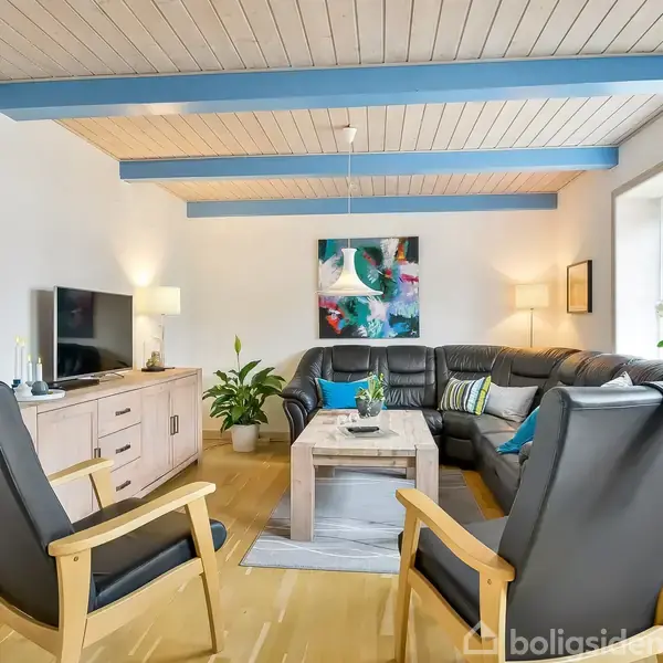 Stue med sort lædersofa og to grå lænestole omkring et sofabord. Fjernsyn på skænk, kunstværk på væggen, planter og lamper skaber en hyggelig atmosfære.