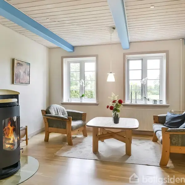 Brændeovn opvarmer hyggelig stue med lyse træmøbler og sofagruppe. Store vinduer lukker dagslys ind, mens blå loftbjælker tilføjer farve. Vase med blomster står på bordet.