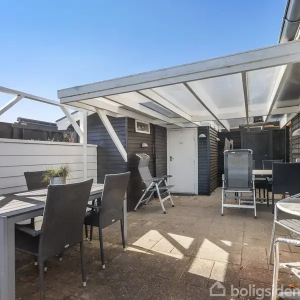Terrasse med sort-hvide møbler under halvtag; bord og stole står på fliser. Omgivet af hvidt plankeværk og sort træbygning. Klar blå himmel ses i baggrunden.