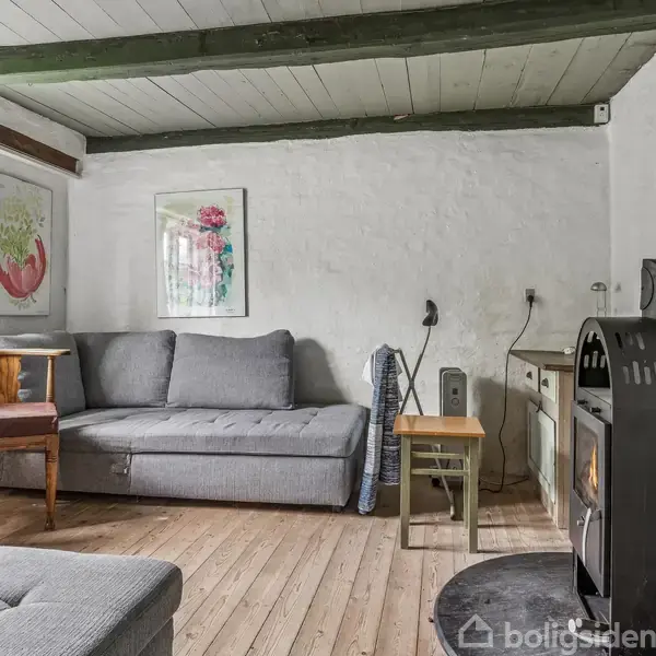 Gråt sofaarrangement i en hyggelig stue med brændeovn; brændeovnen står ved siden af et lille bord og en enkel sofa. Rummet har trægulv, hvide vægge og synlige loftsbjælker.