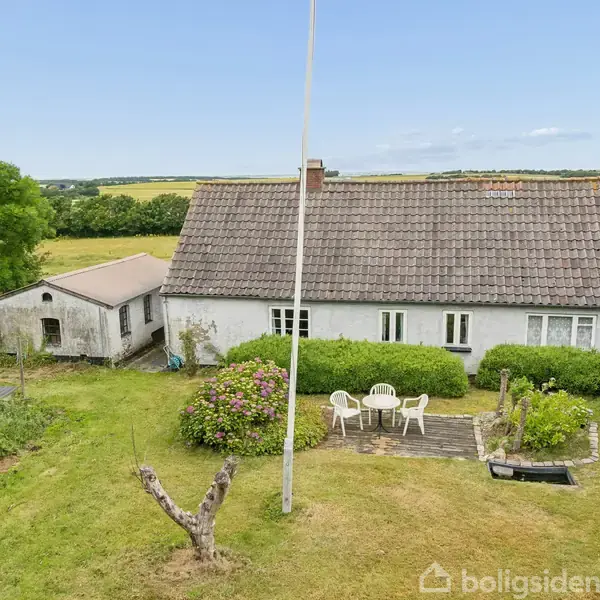 Et hvidt bondehus med rødt tegltag står i en grøn have. Foran huset er et lille terrasseområde med to hvide havestole og bord, omgivet af buske og træer.