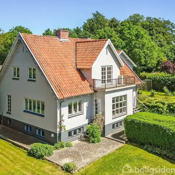 Et klassisk hvidt hus med rødt tegltag står i en velplejet have med hække og plæner. Omkringliggende træer giver en grøn baggrund.