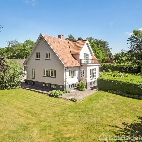 Hvidt hus med rødt tegltag står i en velplejet have med græsplæne og buske. Huset har flere vinduer og en lille altan. Solskin oplyser området.