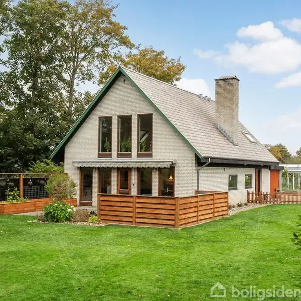 Et hvidt hus med stor terrasse og træhegn placeret midt i en frodig have. Det er omgivet af høje træer under en klar blå himmel.