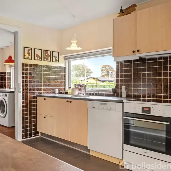 Et køkken med træskabe, sort flisebagvæg og hvide apparater inklusive en opvaskemaskine og komfur. Et vindue viser en have. En dør til venstre fører til et bryggers med vaskemaskine.