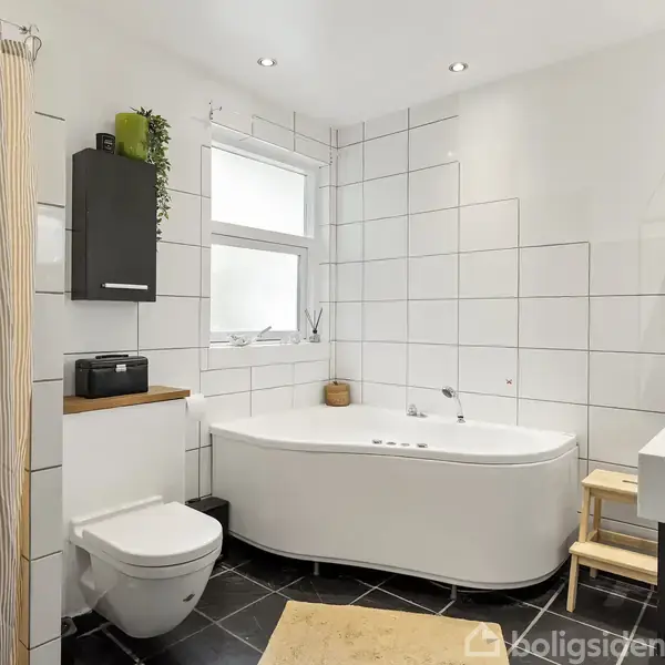 Badeværelse med fritstående badekar i hjørnet. Hængende skab og plante over det væghængte toilet. Håndvask med opbevaring, hylde med dekorative elementer. Fliser på væggene, sort gulv.