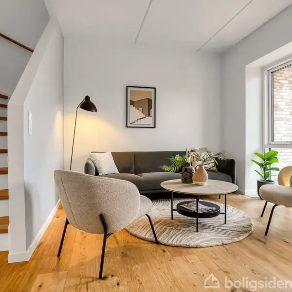 Stue med en mørk sofa og to beige stole omkring et rundt sofabord. En plante står ved vinduet, og en trappe er placeret ved siden af sofaen. Væggene er lyse, og der hænger et billede.