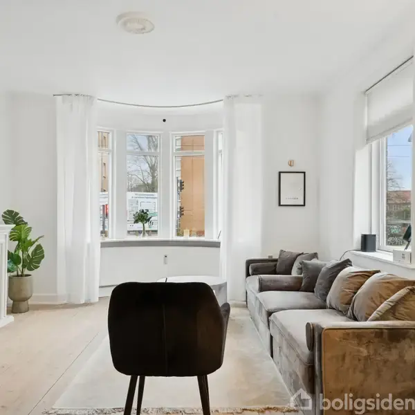 Stue med sofa og stole omkring sofabord, fladskærms-TV på væggen over pejs. Store vinduer med lyse gardiner giver udsigt over bygninger og himmel. Grøn plante i hjørnet.