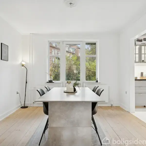 Et spisebord med fire stole står i et lyst rum. Store vinduer i baggrunden viser udsigt til træer. Højre side fører til et køkkenområde.