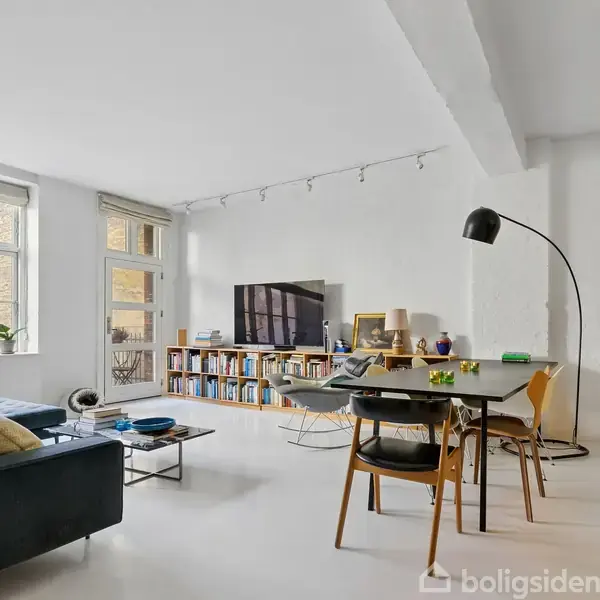 En stue med en sort sofa og et sofabord foran store vinduer. En reol med bøger og en fladskærm på væggen. Et spisebord med stole og en stor gulvlampe ved siden af. På væggen hænger et maleri.