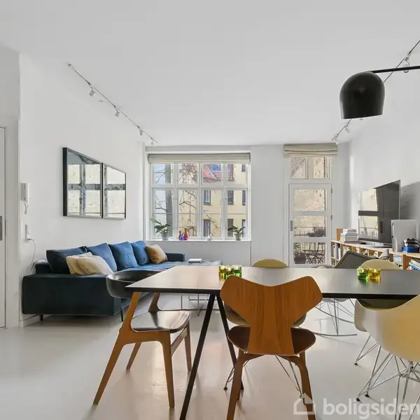 Spisebord omgivet af forskellige stole i lyst værelse. Sofa står mod væggen med billeder, mens vinduer og dør lukker lys ind. Reoler med genstande ses i baggrunden.