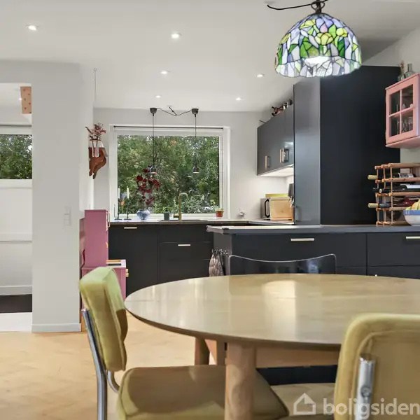 Rundt spisebord med stole står midt i køkkenalrum. Køkkenet i baggrunden har mørke skabe, en stor vindue og lysekrone med farvet glas. Serveicestativ ved siden af farvet vitrineskab.