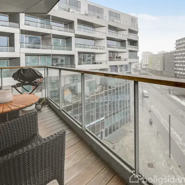 Balkon med trædæk og glasrækværk, indeholdende et rundt bord med to kurvestole og en grill; udsigt til moderne bygninger i et byområde.