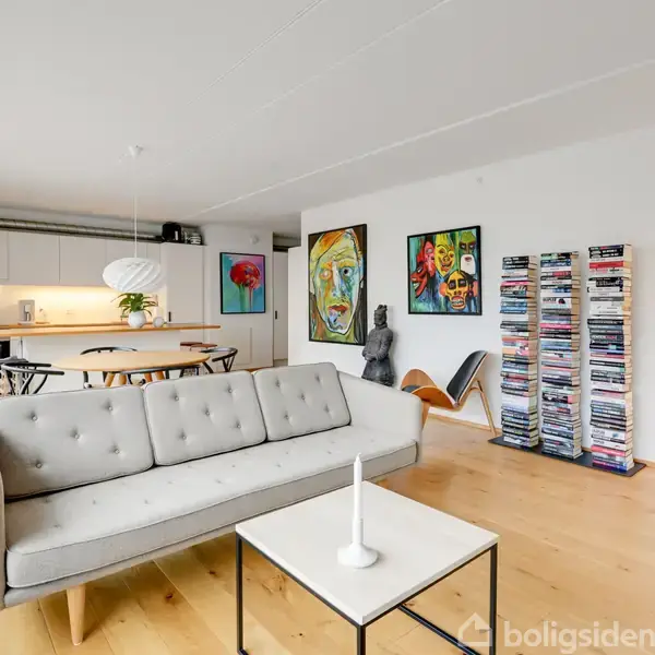 Sofa står i lys stue, omgivet af kunstværker og reolvæg fyldt med magasiner. I baggrunden er der et spiseområde med bord, stole og moderne lampe.