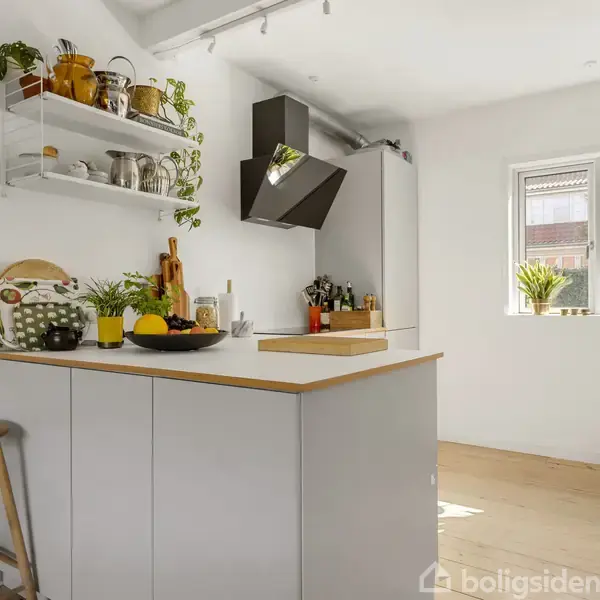 Et køkken med en central ø fyldt med frugt og køkkenredskaber. Hylder med planter og køkkenting hænger over i et lyst rum med en stor vindueskarm dekoreret med grønne planter.