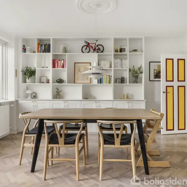 Spisebord med seks stole står i et lyst rum. Reolen i baggrunden er fyldt med bøger og planter. En cykel pynter ovenpå, mens vinduer giver udsigt over grønt område.