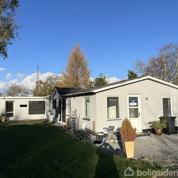 Et hvidt hus med fladt tag står stille i en have med grønt græs og træer. Et skilt i et vindue siger "Til Salg".