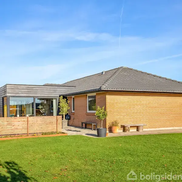 Et moderne enfamiliehus står på en velplejet græsplæne med blå himmel over. En terrasse med store vinduer findes til venstre, og omkransende hæk fuldender haven.