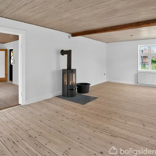 Stue med en sort brændeovn i hjørnet, omgivet af trægulv og hvidmalede vægge. Et åbent dørparti fører til et tilstødende rum. En radiator under vinduet.